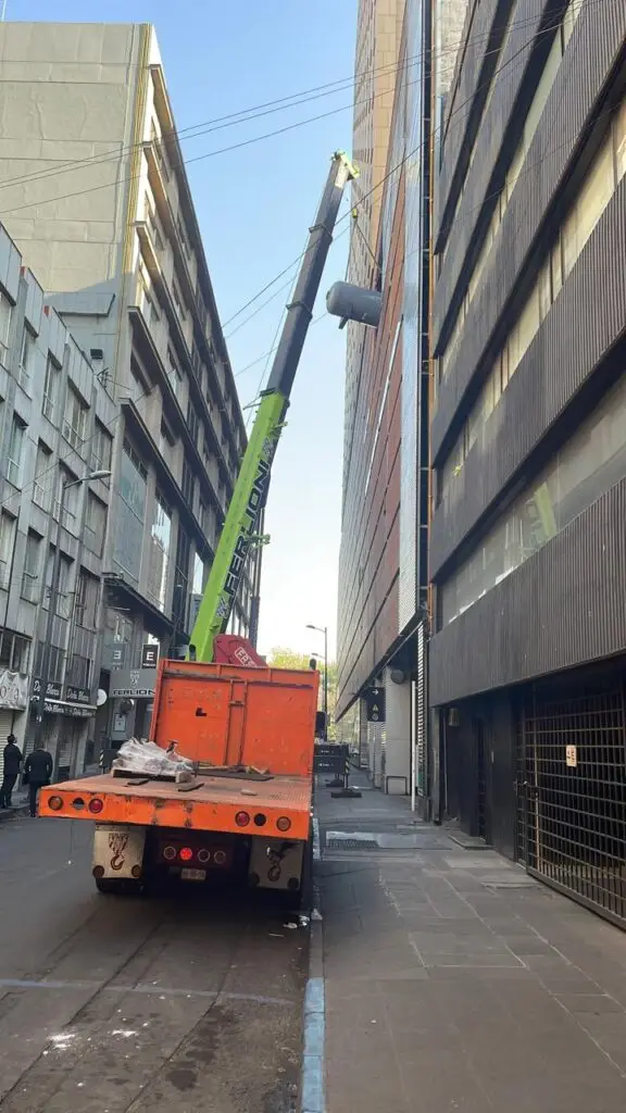Grúa de gran tamaño elevando tanque estacionario de gas frente a edificio alto en el centro de la Ciudad de México, operado desde camión de maniobras marca Ferlion.